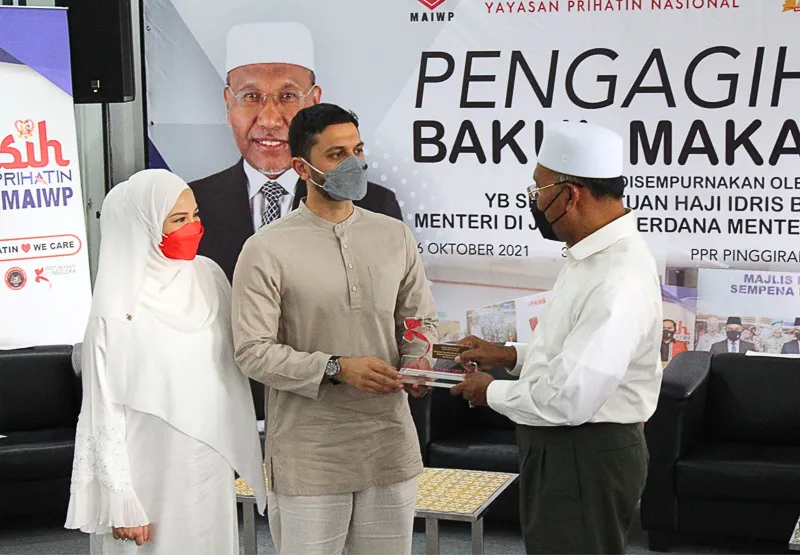 Three people wearing face masks and traditional Malay attire, with one man handing an award or plaque to another man, in front of a banner about a food distribution event.