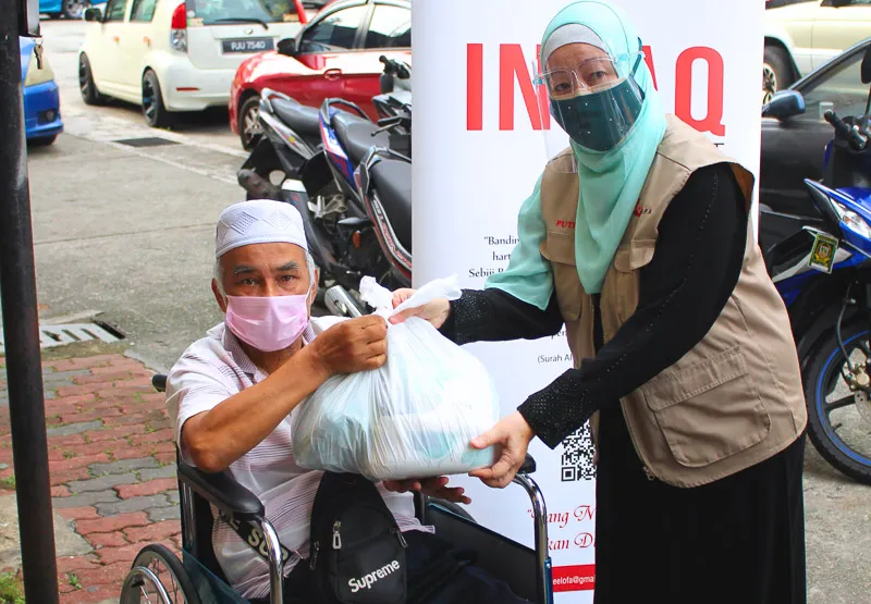Woman wearing a hijab, face shield, and mask handing a plastic bag of supplies to a man in a wheelchair wearing a white cap and mask.
