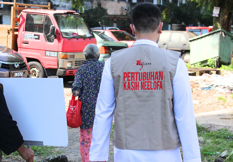 Man wearing a vest with 'Pertubuhan Kasih Neelofa' walking behind an elderly woman carrying a red bag, near parked vehicles.