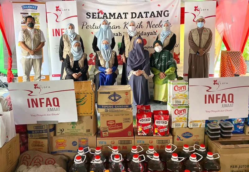 A group of masked people posing behind stacked food and beverage donations at a charity event with banners that read 'INFAQ Jumaat' and 'Selamat Datang Pertubuhan Kasih Neeba'.