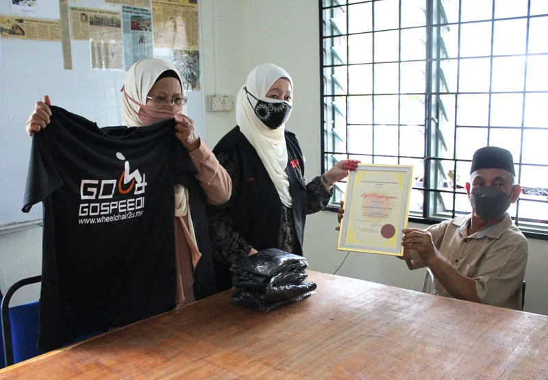 Two women wearing headscarves and face masks present a certificate and a stack of black GOSPEED T-shirts to a seated man in a cap and face mask beside a wooden table.