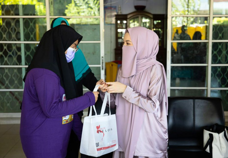 Two Muslim women wearing hijabs exchanging a white shopping bag indoors, with a third woman partially visible in the background.