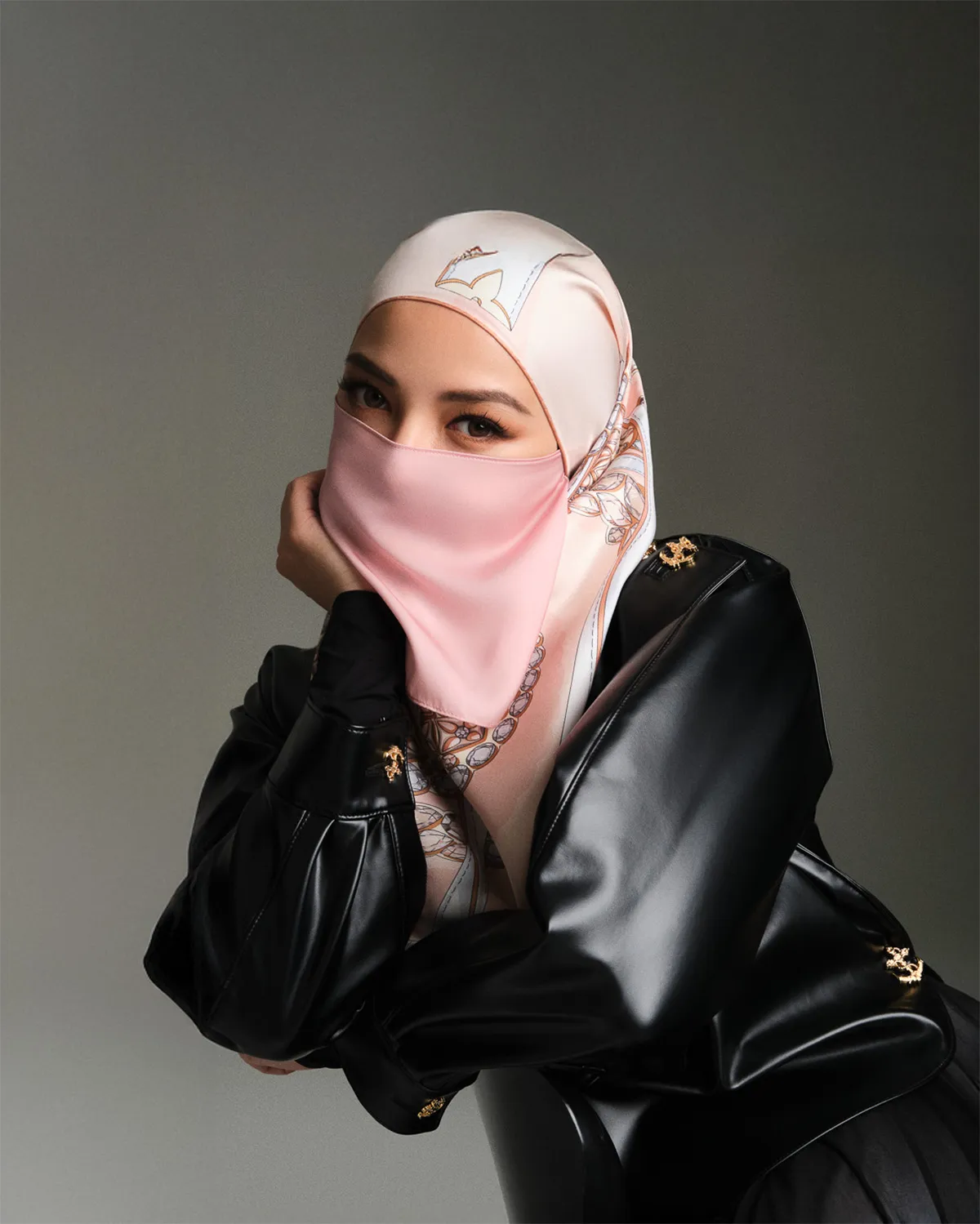 Woman wearing a pink patterned silk hijab and matching face covering, dressed in a black leather jacket, posing with her hand near her face against a gray background.