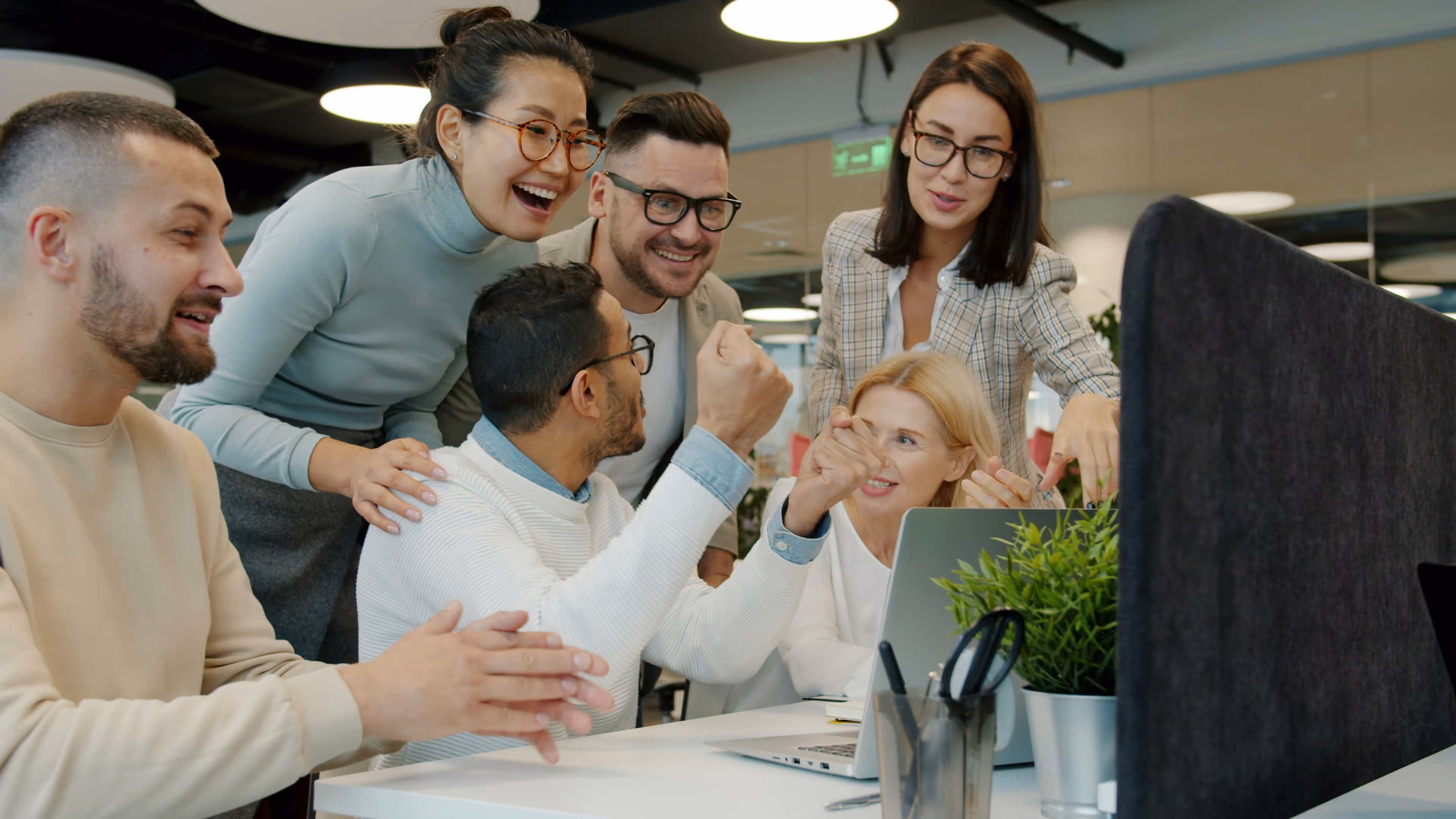 Grupo diverso de compañeros de trabajo celebrando frente a una computadora portátil en una oficina moderna.
