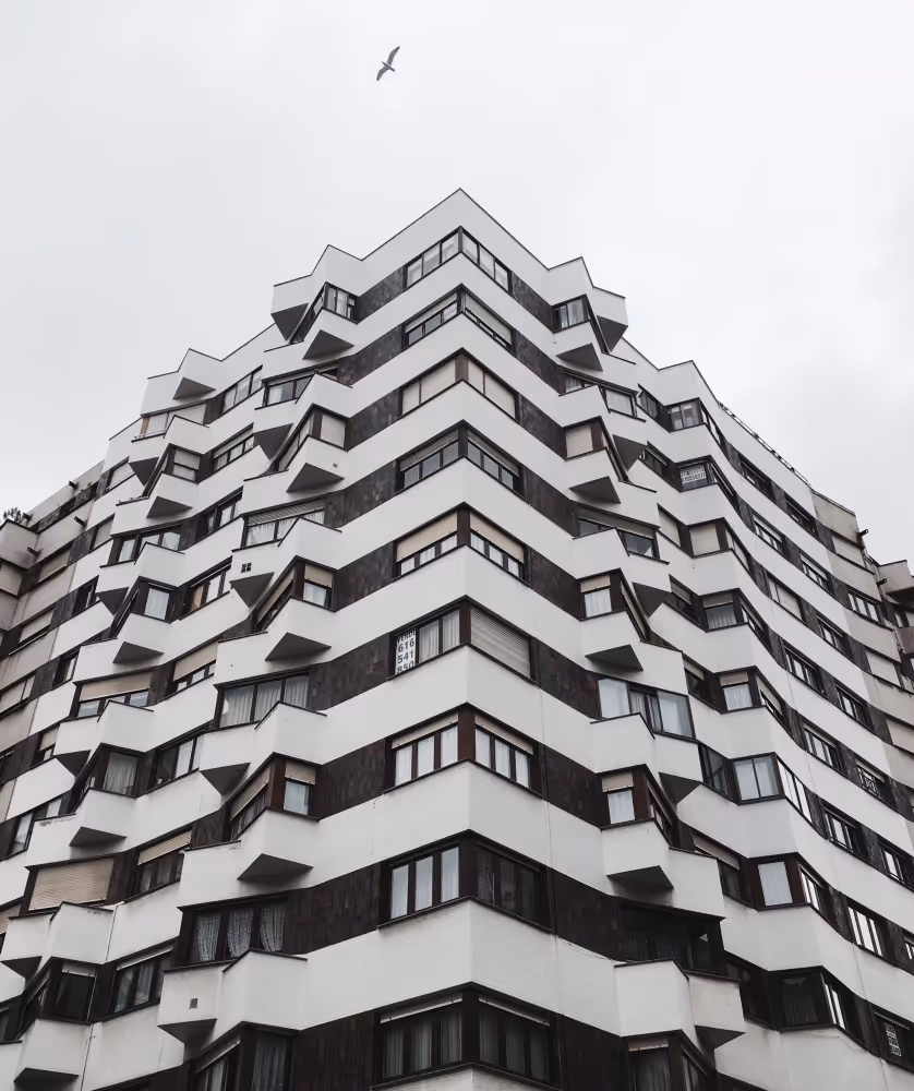 Blick nach oben auf ein mehrstöckiges modernes Wohngebäude mit einem Vogel am bewölkten Himmel.