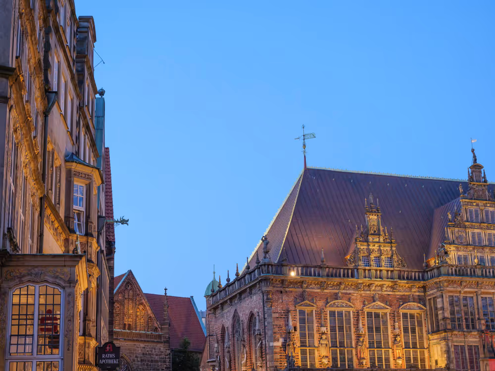 Luftaufnahme eines historischen Gebäudes mit rotem Backstein und grünem Turmdach in einer Stadt mit weiteren Gebäuden und Bäumen.