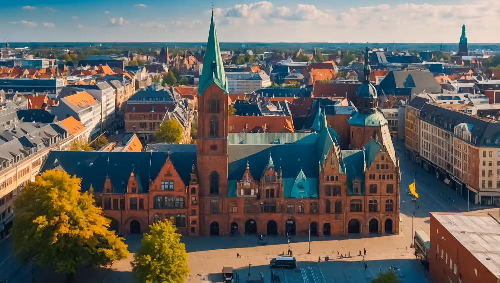 Luftaufnahme eines historischen Gebäudes mit rotem Backstein und grünem Turmdach in einer Stadt mit weiteren Gebäuden und Bäumen.