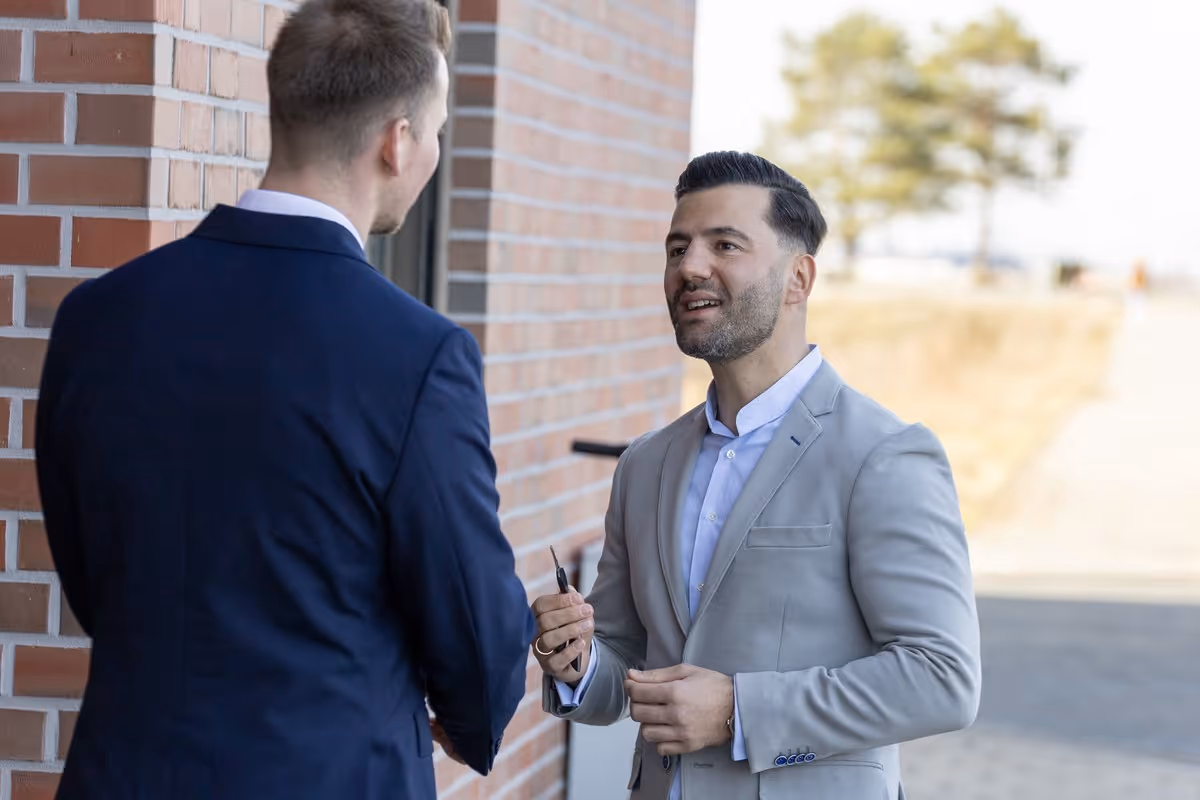 Zwei Männer in Anzügen sprechen draußen neben einem Backsteingebäude, einer hält einen Schlüssel.
