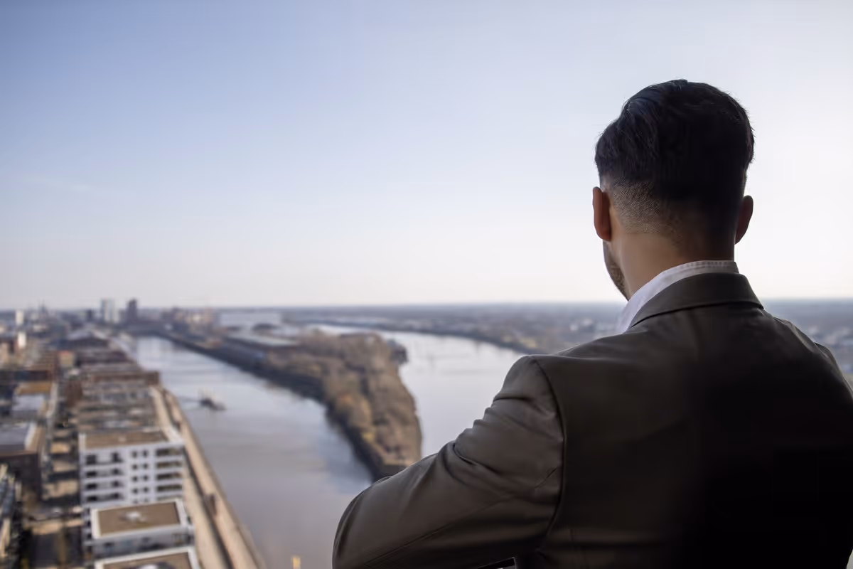 Ein Mann in Anzug blickt von einem hohen Gebäude auf einen Fluss und eine Stadtlandschaft.