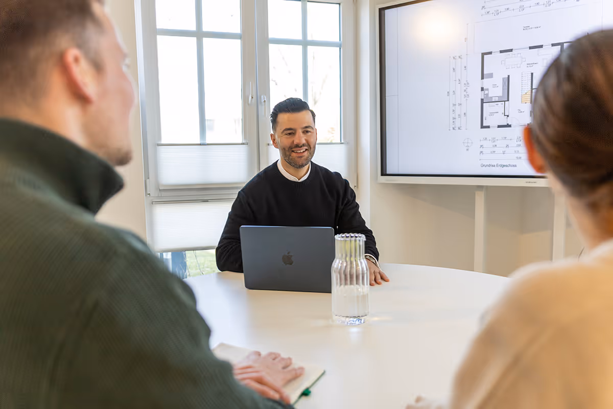 Drei Personen bei einer Besprechung in einem hellen Raum, mit einem Laptop, einem Glas wasser und einem Grundriss auf einem Bildschirm.