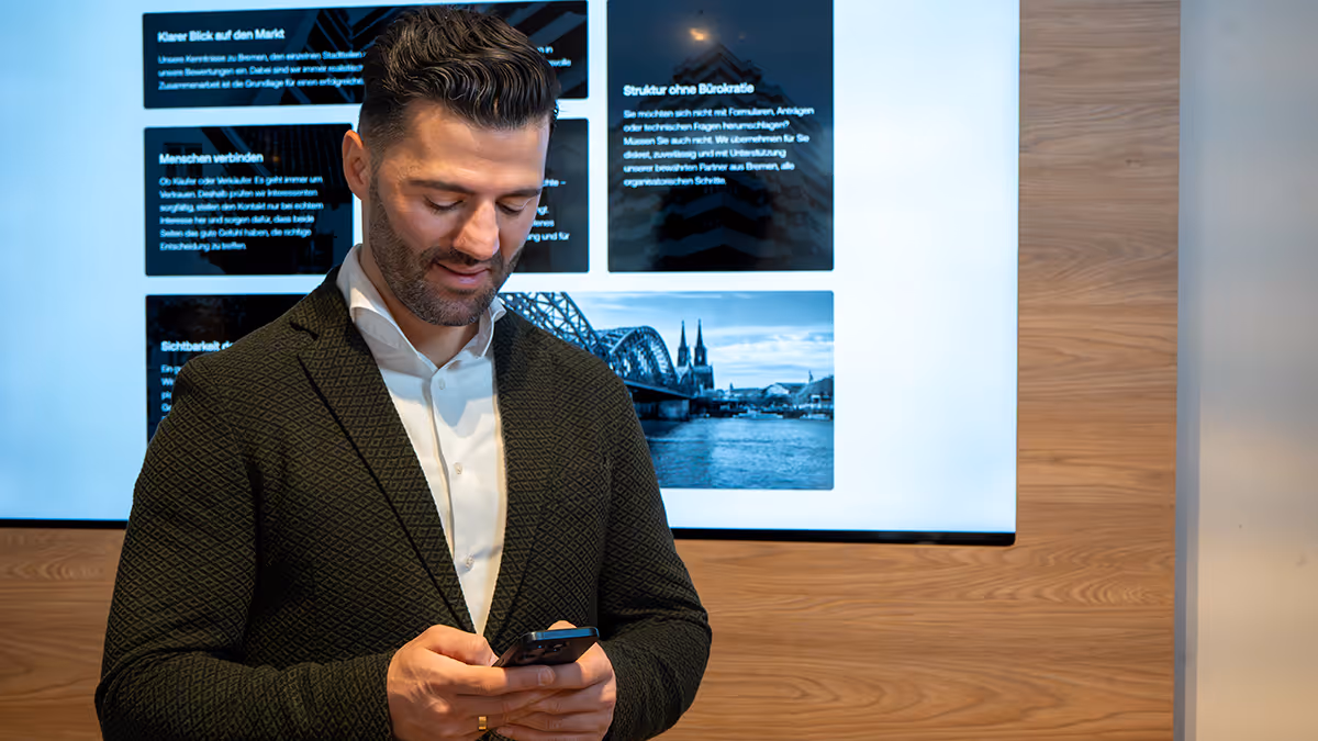 Mann mit Anzug blickt auf sein Handy vor einem Bildschirm mit Text und einem Bild der Kölner Dom und der Hohenzollernbrücke.