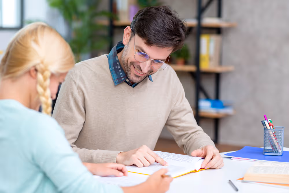 Mann mit Brille erklärt einem Kind mit blonden Zöpfen einen Text aus einem aufgeschlagenen Buch am Tisch.