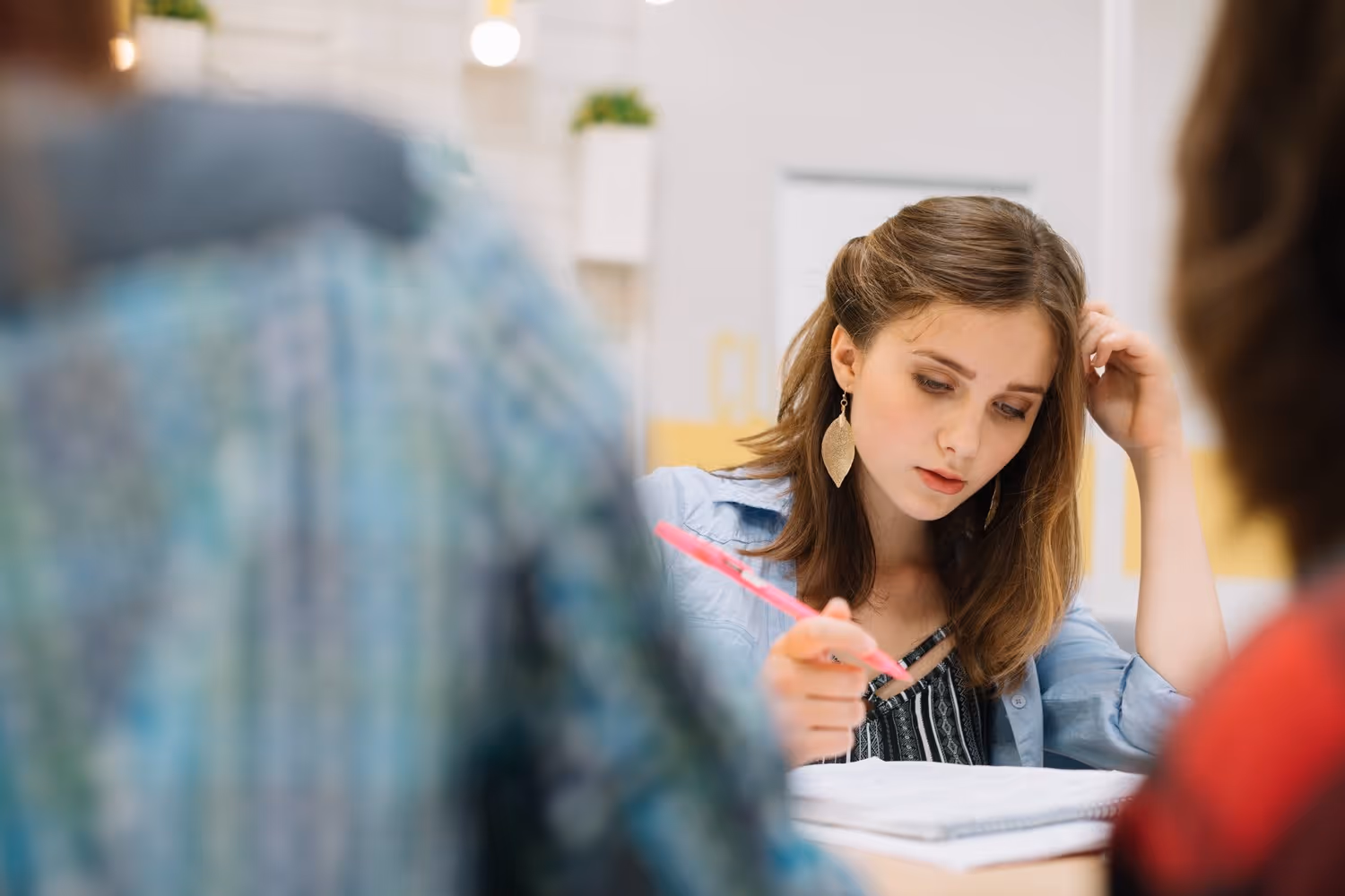 Junge Frau mit braunen Haaren schreibt konzentriert mit einem pinken Stift in einem Notizbuch in einem Klassenzimmer.