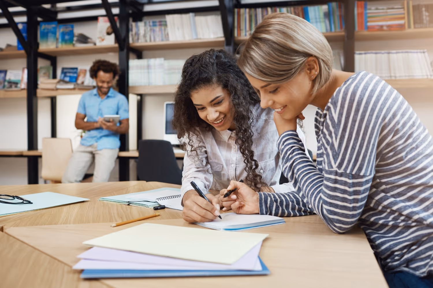 Zwei Frauen lernen gemeinsam an einem Tisch mit Notizbüchern und Ordnern, während ein Mann im Hintergrund ein Tablet benutzt.