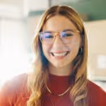 Junge Frau mit Brille und langen blonden Haaren, die lächelnd in die Kamera schaut.