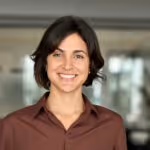 Lächelnde Frau mit kurzen dunklen Haaren und braunem Hemd vor unscharfem Bürohintergrund.