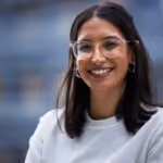 Junge Frau mit schwarzen Haaren, Brille und weißem Pullover lächelt vor unscharfem Hintergrund.