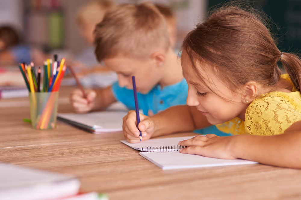 Grundschulkinder konzentrieren sich beim Schreiben in Notizbüchern an einem Tisch mit bunten Buntstiften.