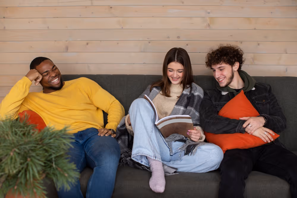 Drei junge Erwachsene sitzen entspannt auf einem Sofa, eine Frau liest aus einem Buch, die beiden Männer lachen dabei.