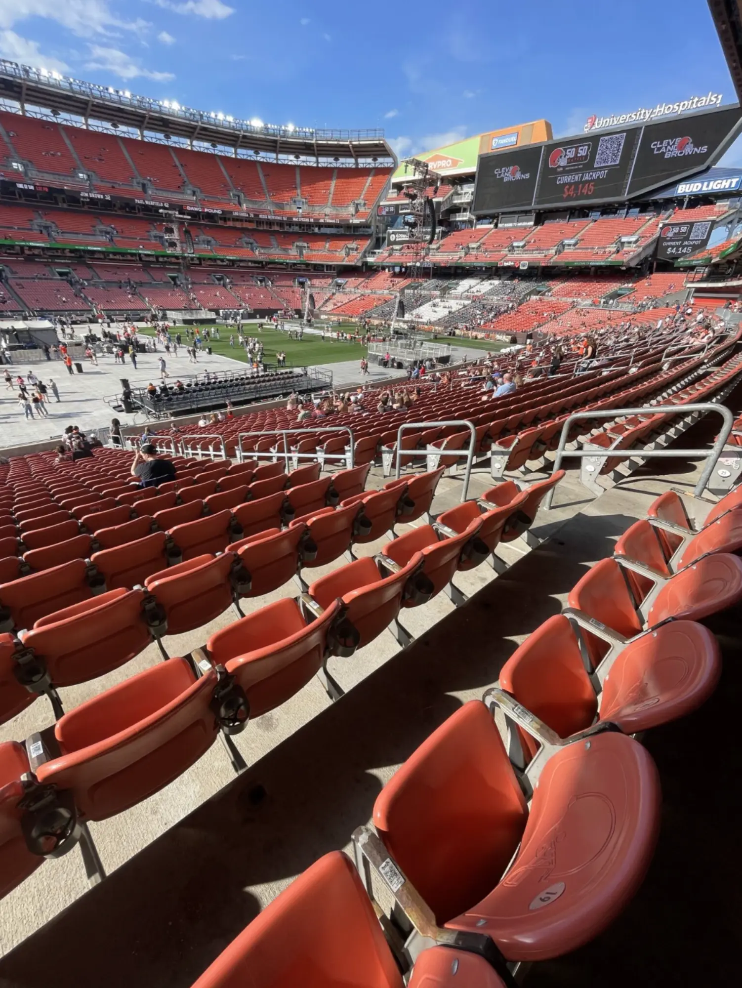Wide view of empty orange stadium seats and a partially filled football field with people preparing under a clear blue sky.