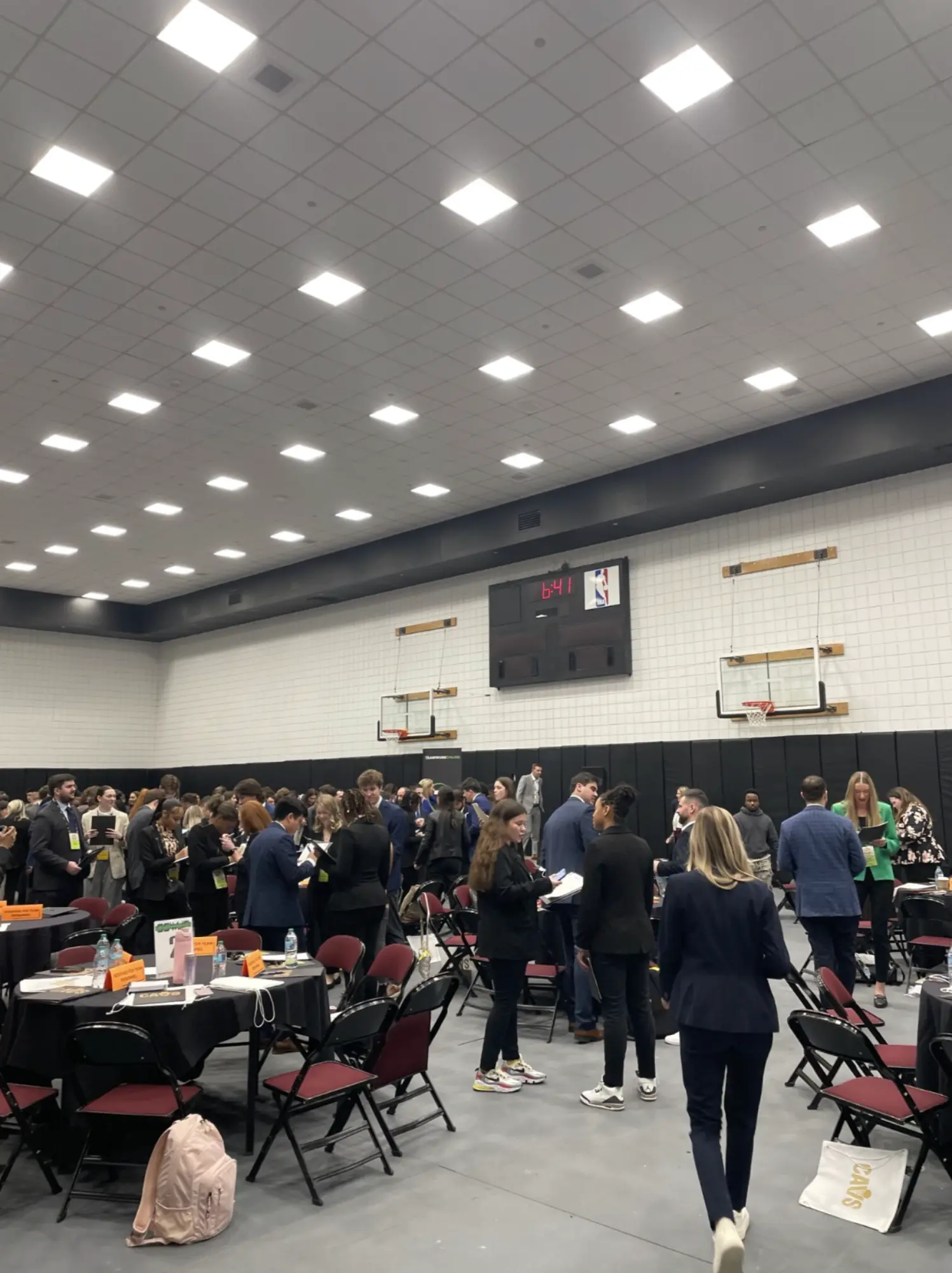 Group of people networking and talking in a gymnasium with folding chairs and tables, and a basketball scoreboard showing 6:41.