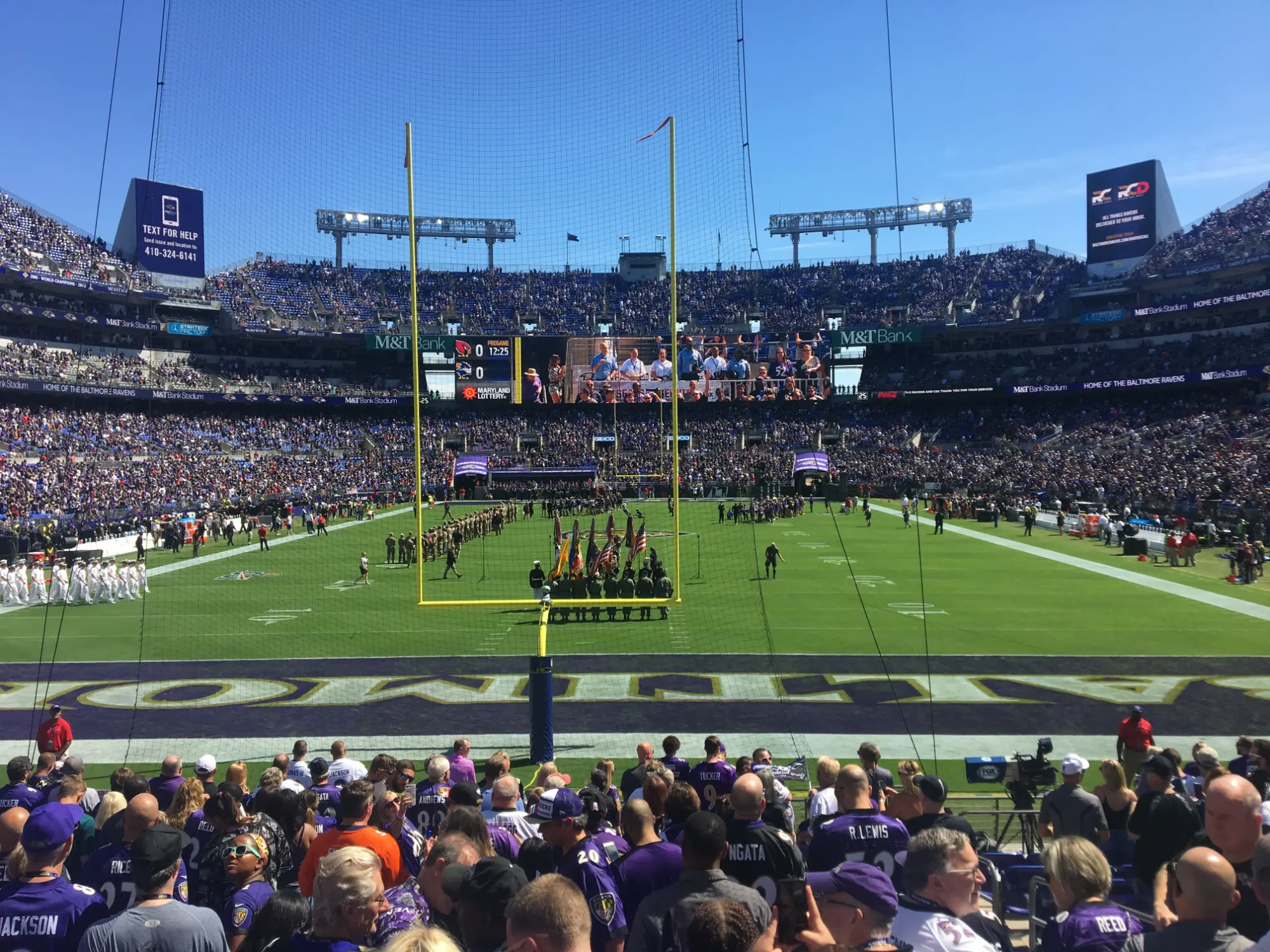Crowded M&T Bank Stadium with fans watching a pregame ceremony featuring a color guard on the football field under clear blue sky.