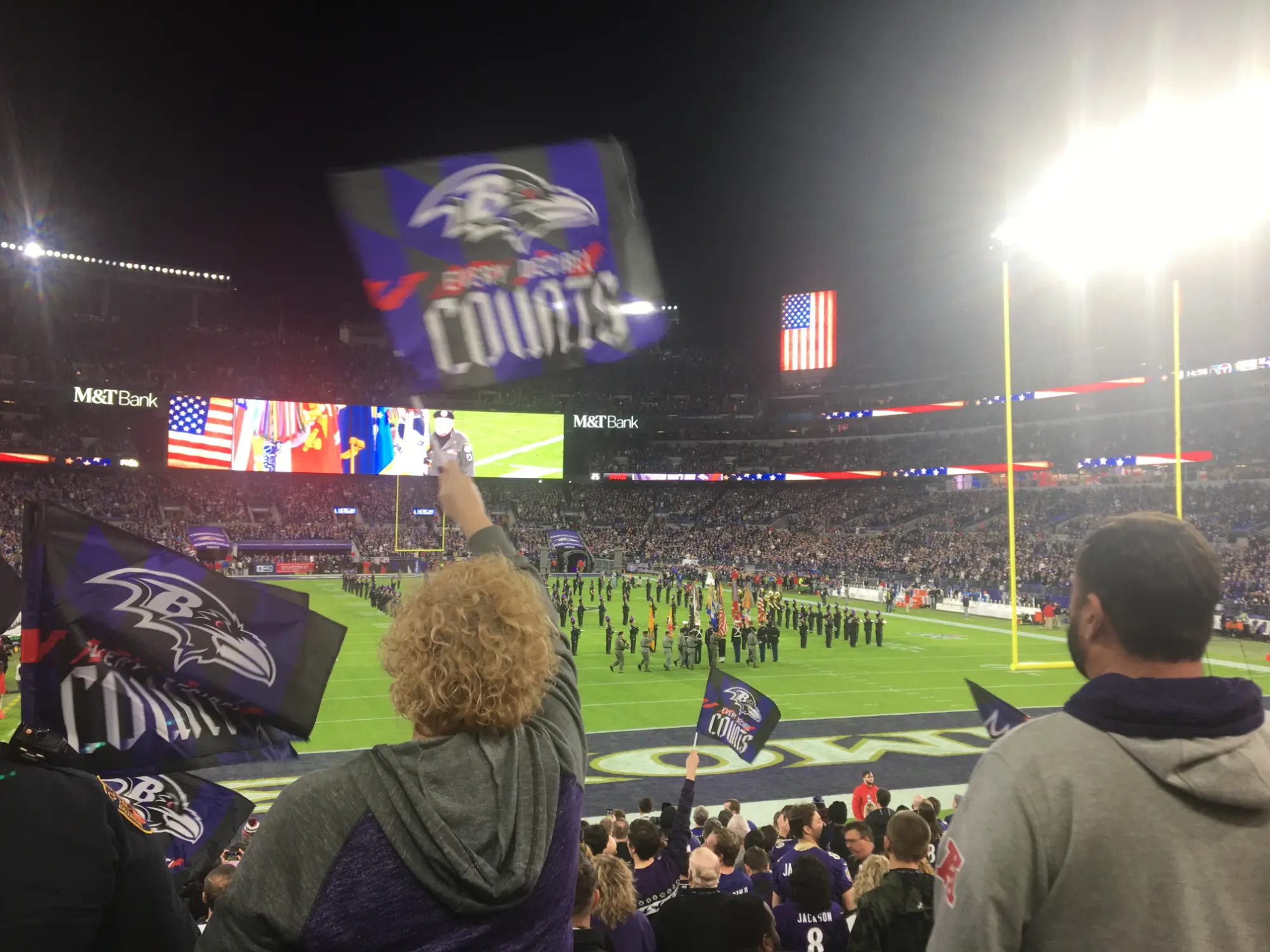 Fans waving Baltimore Ravens flags in a crowded stadium during a nighttime football game with the field and American flags visible.