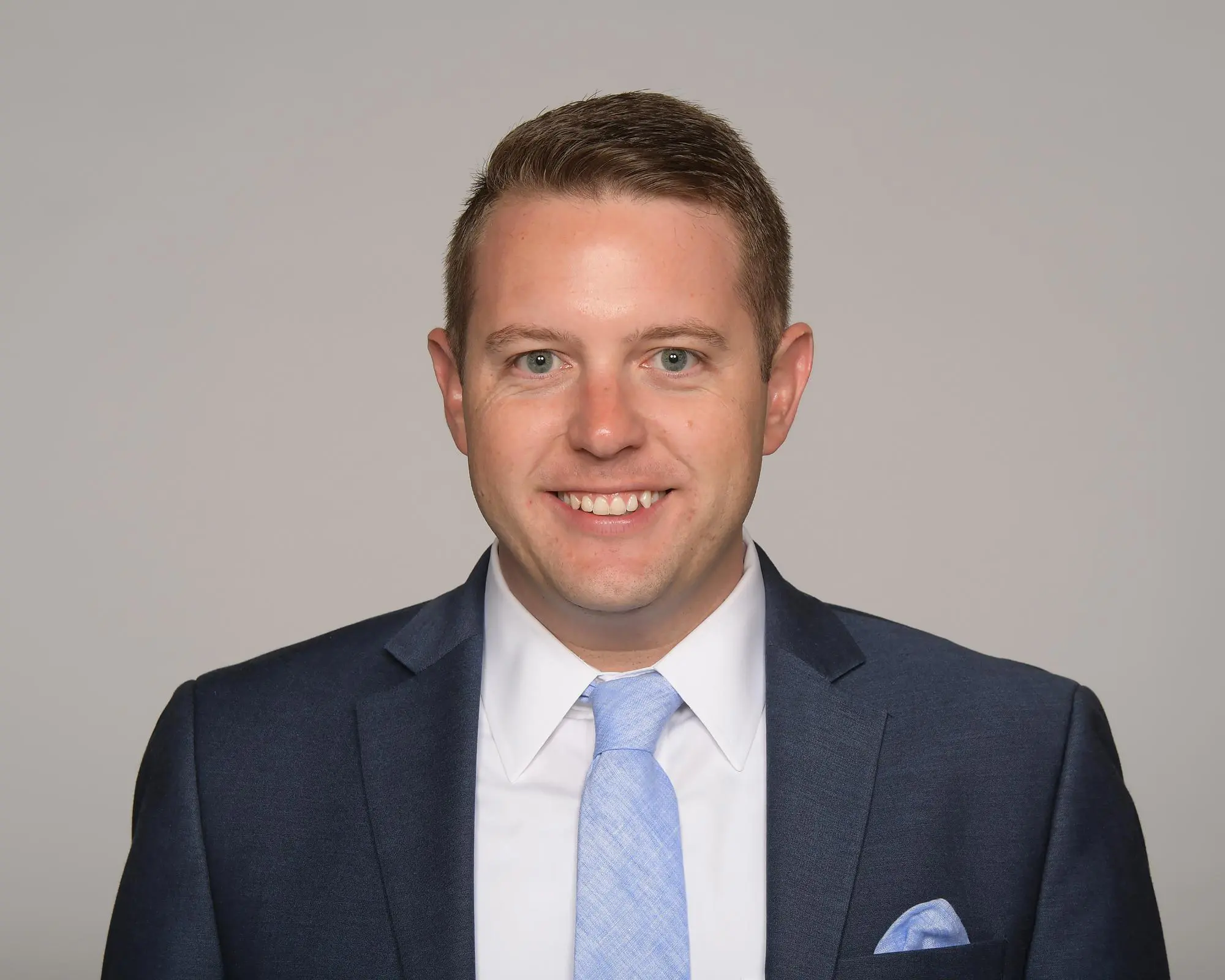 Smiling man with short brown hair wearing a navy blue suit, white shirt, and light blue tie, against a gray background.