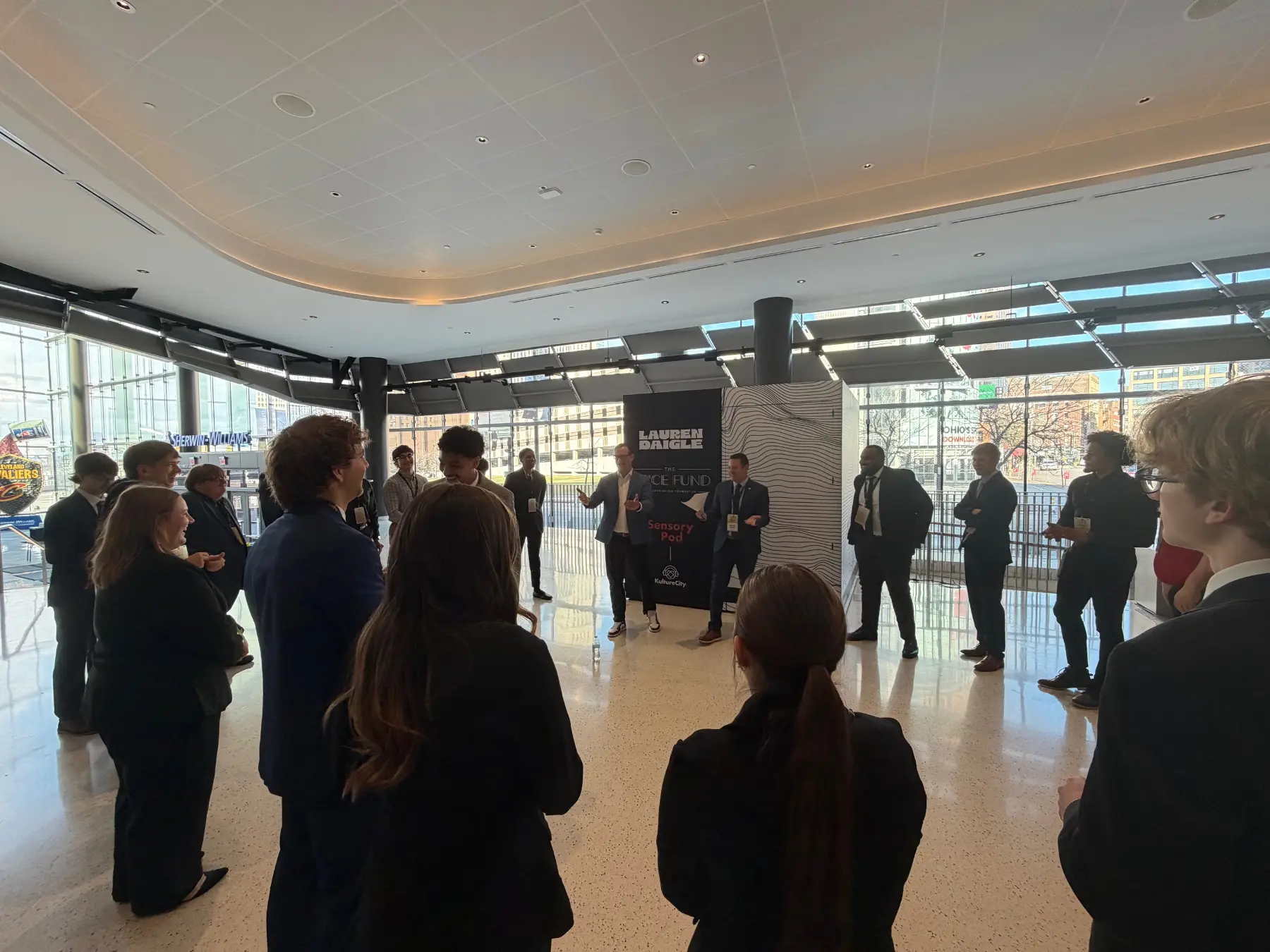 Group of people standing in a spacious modern lobby, listening to two men speaking near a display labeled 'Lauren Daigle Sensory Pod.'