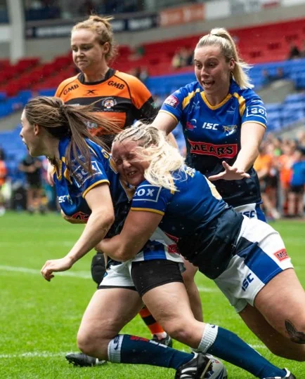 Women playing rugby with one player in orange and three in blue jerseys engaged in a tackle on a grass field.