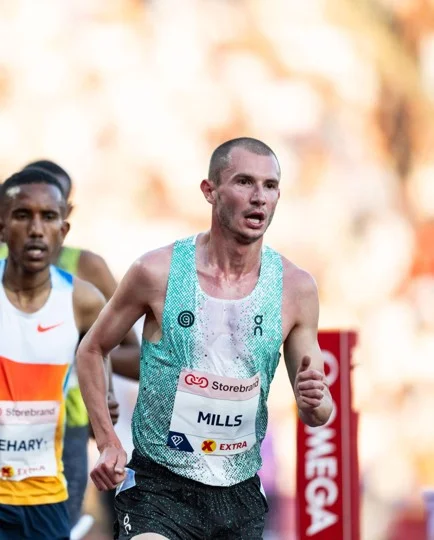 Male runner wearing a green and white singlet with bib labeled 'MILLS' competing in a race.