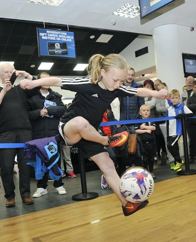 Young girl in black sportswear skillfully controlling a soccer ball mid-air indoors with an audience watching behind a blue barrier.