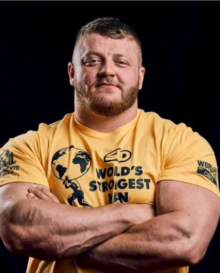 Muscular man with short hair and beard wearing a yellow 'World's Strongest Man' t-shirt with arms crossed.
