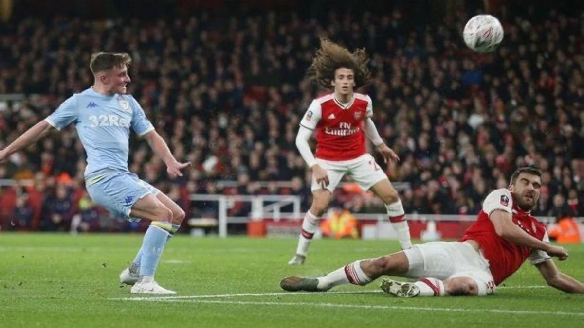 Soccer player in light blue jersey kicking the ball while two players in red and white jerseys try to block it on the field.
