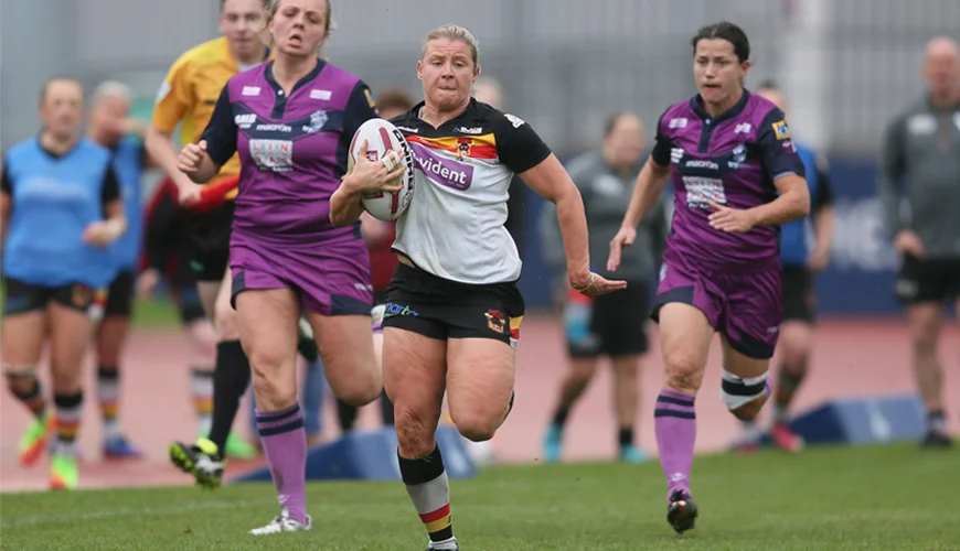 Female rugby player in white and black jersey running with ball while two opponents in purple jerseys chase her on a grass field.