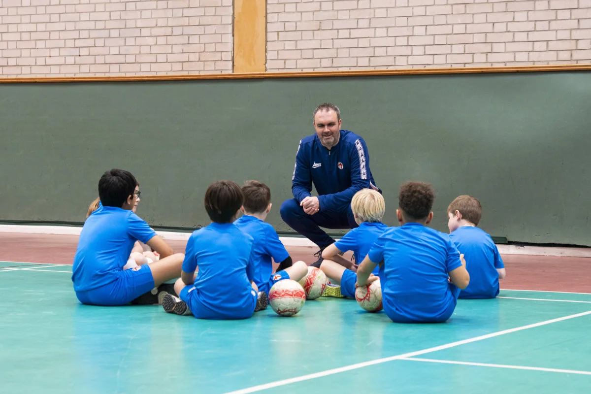 Children in blue sports uniforms sitting on a gym floor while a coach kneels and talks to them.