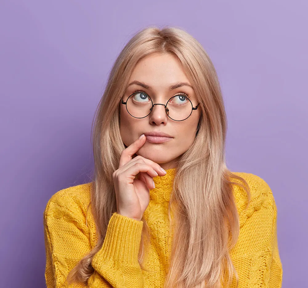 Junge blonde Frau mit runden Brillengläsern und gelbem Pullover, die nachdenklich nach oben schaut.