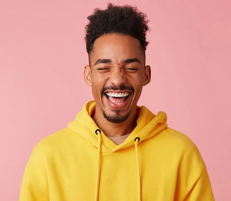 Junger Mann mit gelber Kapuzenjacke lacht mit geschlossenen Augen vor rosa Hintergrund.