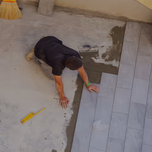 Ein Fliesenleger kniet auf dem Boden und verlegt rechteckige graue Bodenfliesen auf frisch aufgetragenen Kleber.