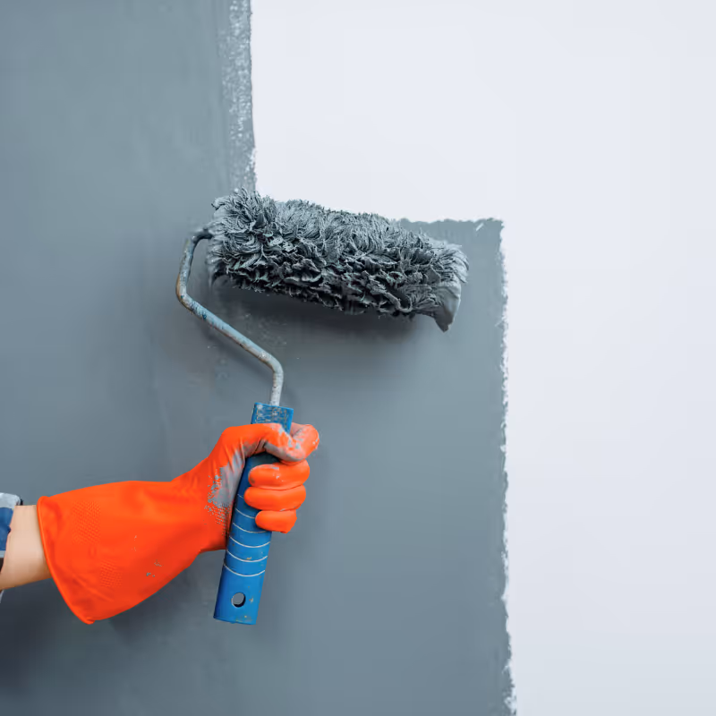 Hand mit orangefarbener Arbeitshandschuh hält Farbroller und streicht graue Farbe auf weiße Wand.