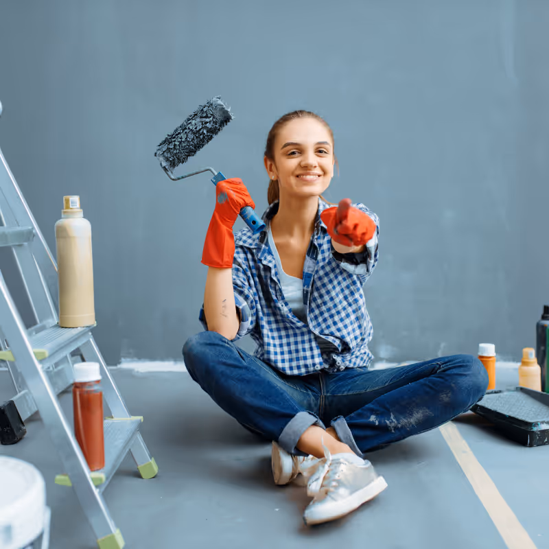 Lächende Frau in kariertem Hemd und roten Handschuhen sitzt mit ausgebreiteten Beinen auf dem Boden und hält Farbroller in der Hand.
