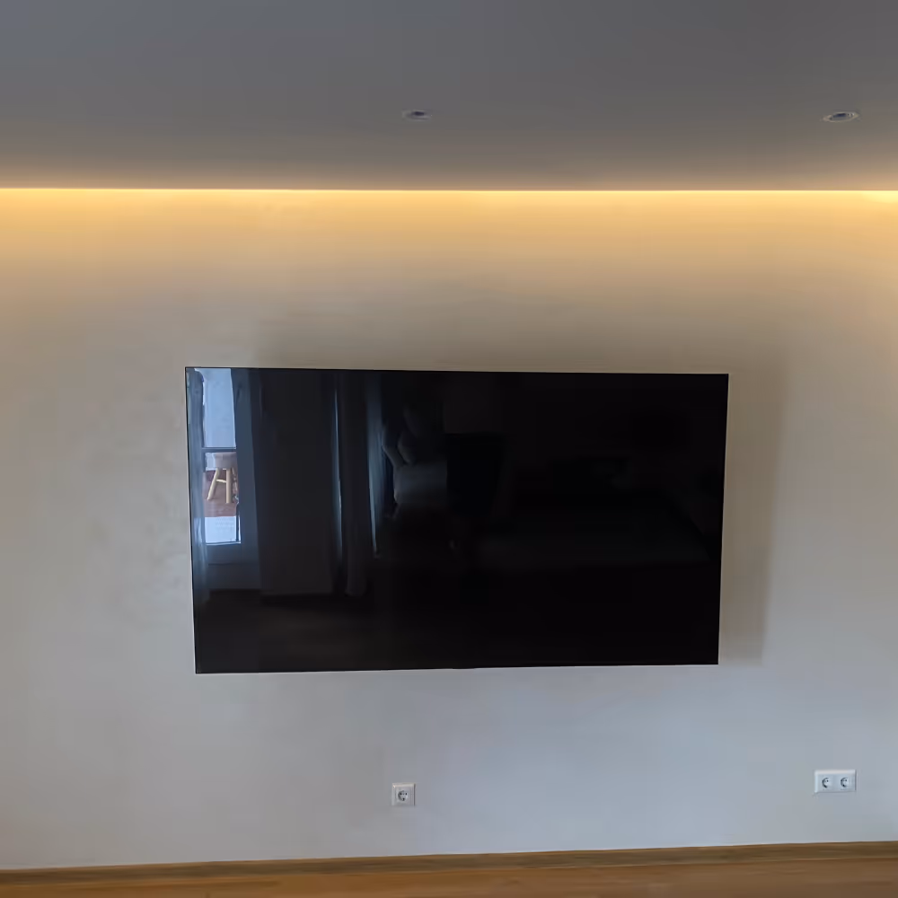 An einem weißen Wandhintergrund hängt ein flacher, schwarzer Fernseher, darüber verläuft eine warme LED-Lichtleiste, unter dem Fernseher befinden sich zwei Steckdosen.