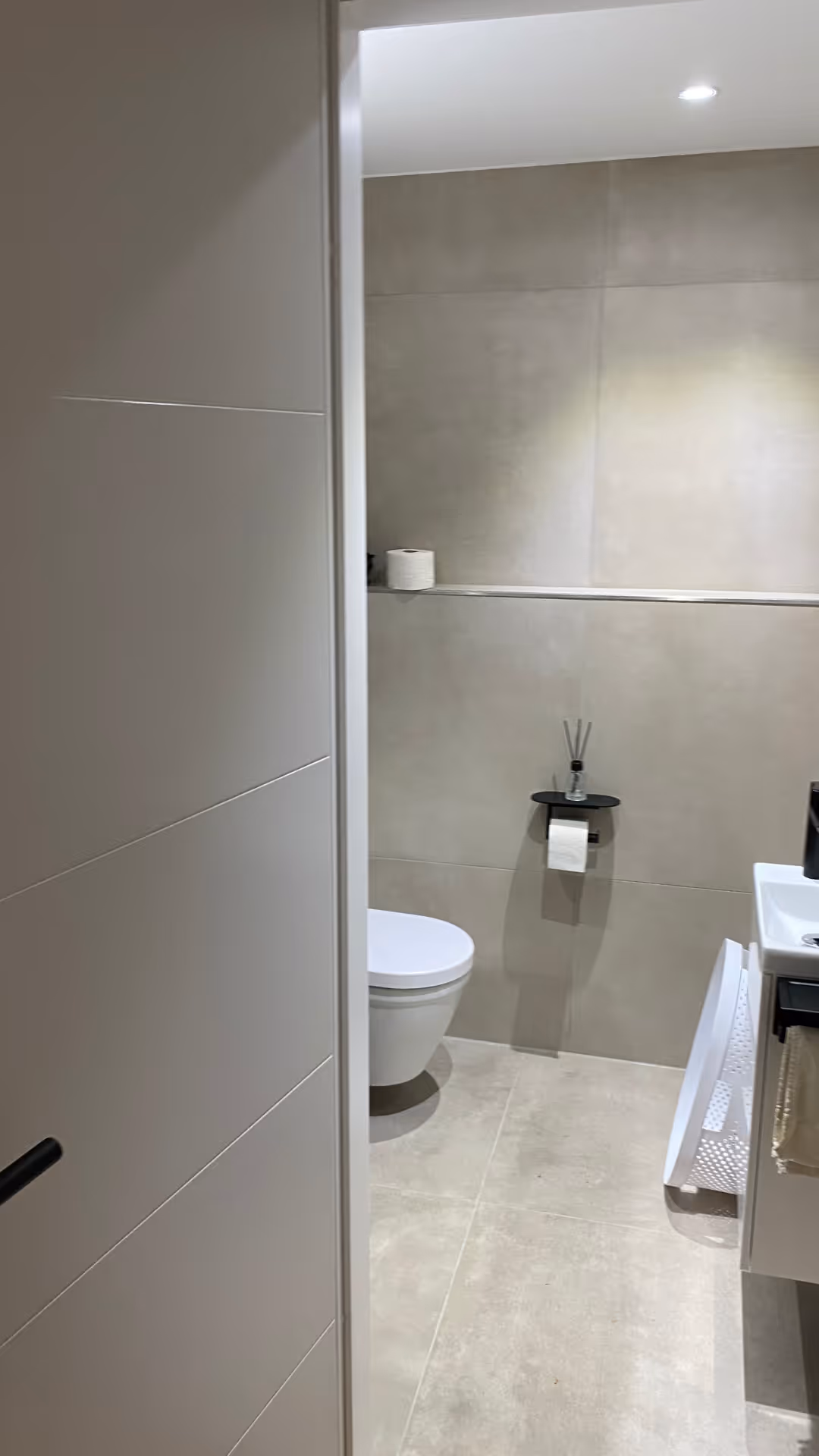 Moderner, sauberer Badezimmereingang mit grauen Fliesen, einer Wandtoilette, an der Wand befestigtem Toilettenpapierhalter und einem weißem Wäschekorb neben dem Waschbecken.