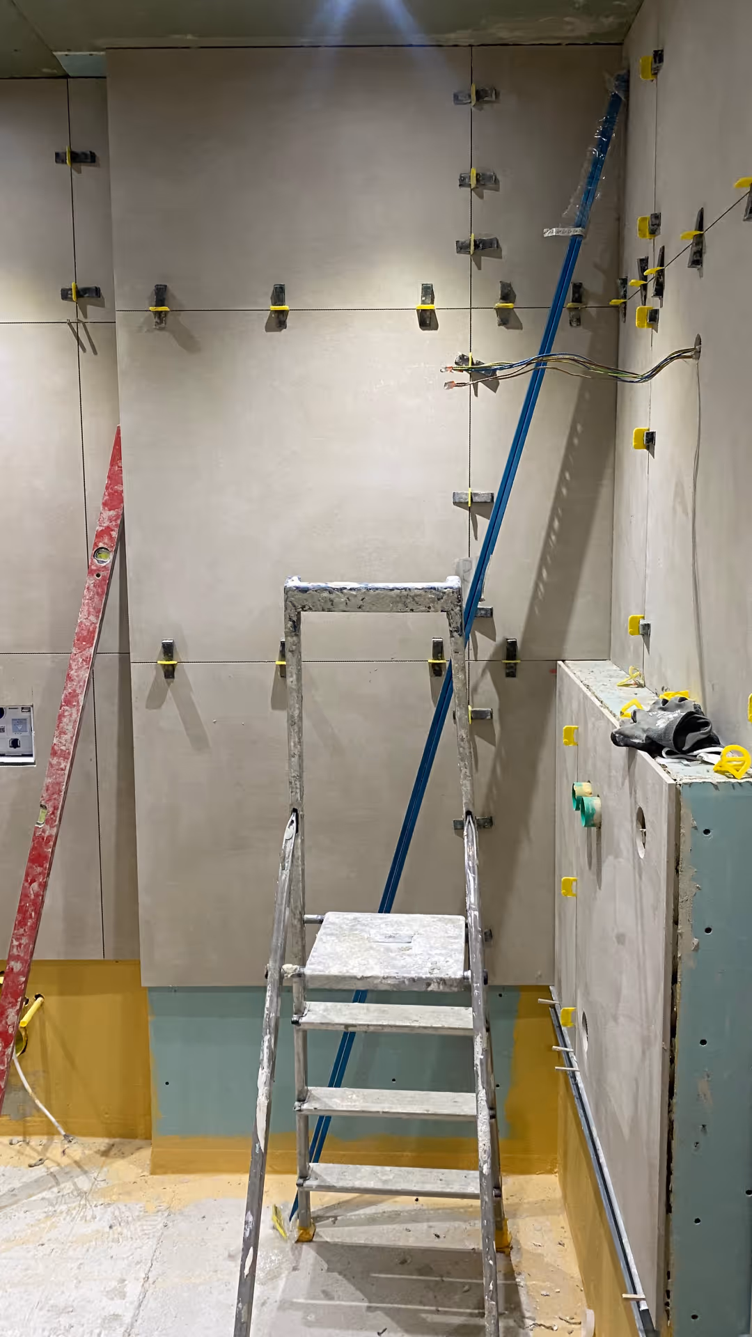 Baustelle eines Badezimmers mit höhenverstellbarem Gerüst, roten Leiter und grauen Wandfliesen in Arbeit.