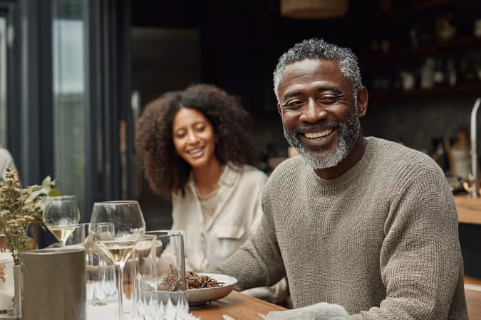 Smiling man with gray beard and sweater sitting at a dining table with a woman and wine glasses.