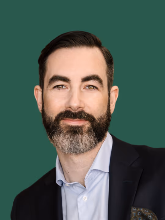 Portrait of a man with dark hair and a salt-and-pepper beard, wearing a light blue shirt and dark blazer against a green background.