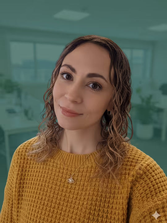 Woman with wavy brown hair wearing a mustard yellow knit sweater and a small pendant necklace, smiling slightly in an office setting.