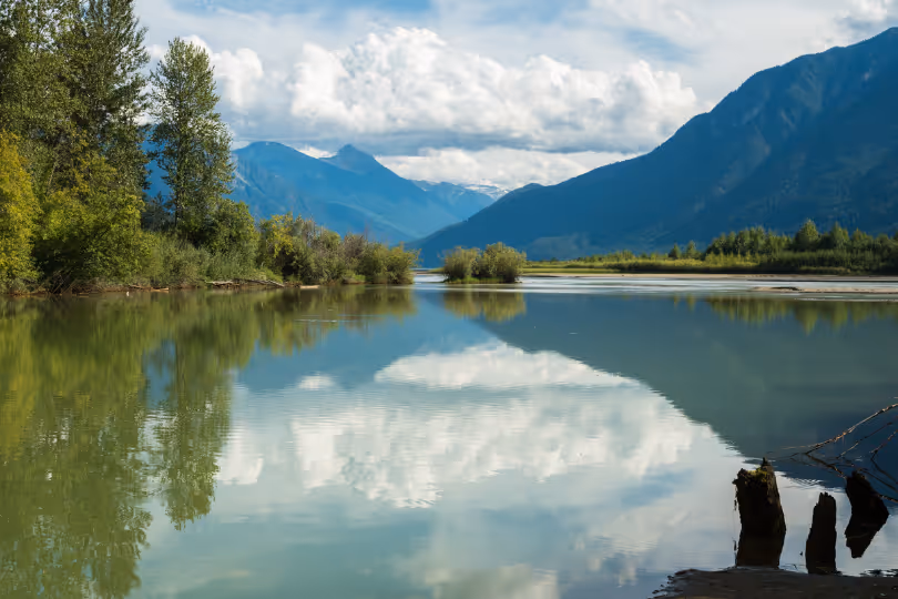 Calm river reflecting trees, white clouds, and mountains under a blue sky.