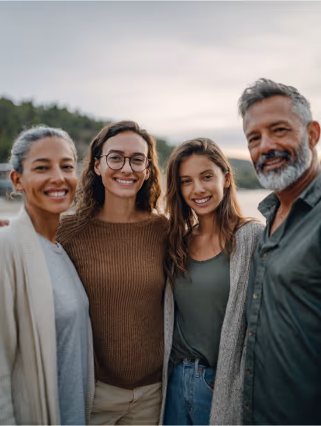 Smiling family of four standing close together outdoors with trees and water in the background.