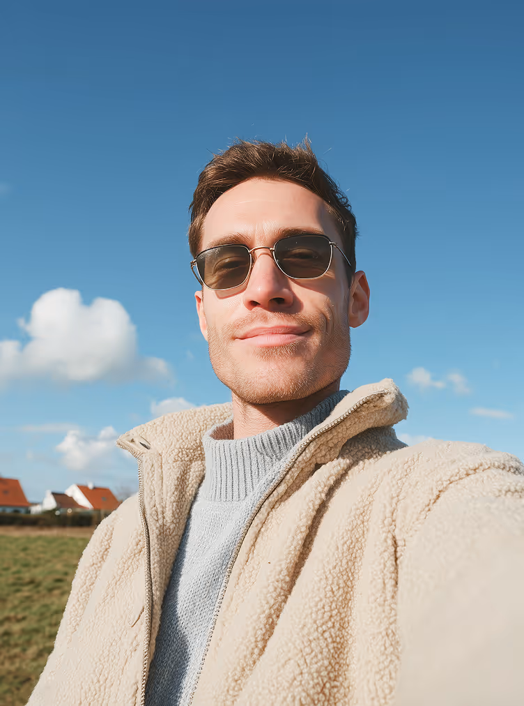 Smiling man wearing sunglasses and a beige fleece jacket taking a selfie outdoors under a blue sky.